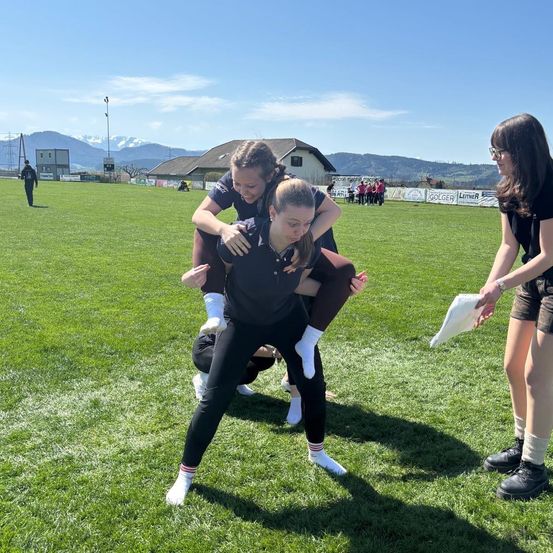 Drei Frauen sind auf einem grünen Feld. Eine Frau hält eine andere Frau auf ihren Schultern. Eine Frau neben ihnen hält ein Papier. In der Ferne gibt es Berge und Gebäude.