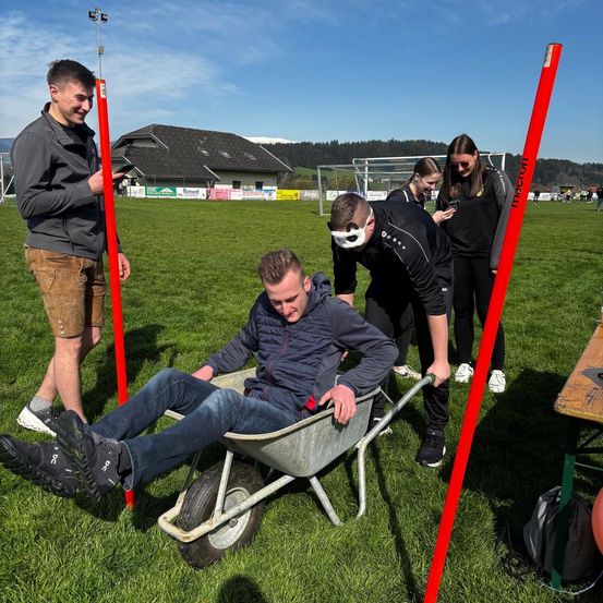 Ein Mann wird von zwei anderen in einer Schubkarre auf einem Rasenfeld geschoben, mit einem Gebäude im Hintergrund.