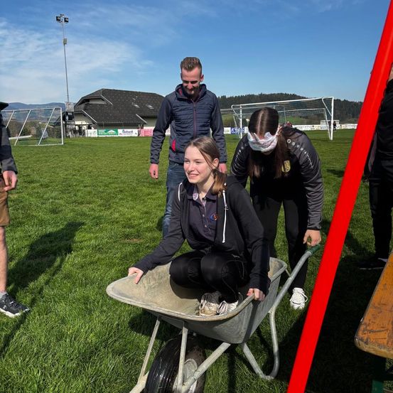 Ein Mädchen sitzt in einer Schubkarre auf einem Rasenfeld, eine Frau hält den Griff. Ein Mann steht hinter ihnen und lächelt. Ein Fußballtor und ein Haus sind im Hintergrund.