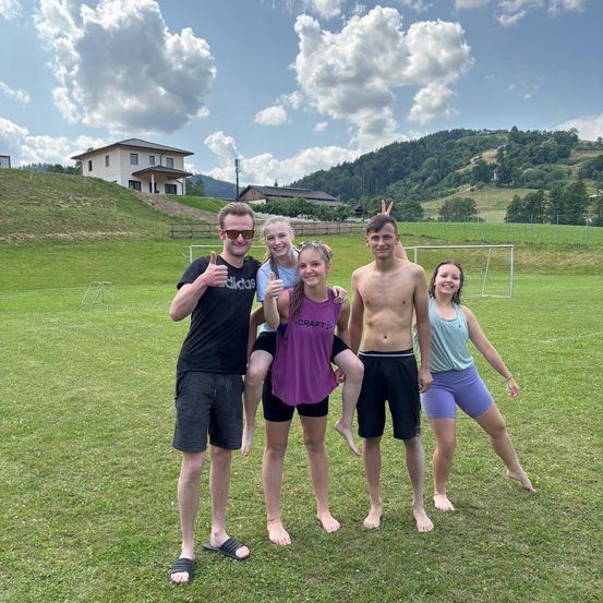 Fünf Menschen posieren für ein Foto auf einem Rasenfeld mit einem Fußballtor. Zwei Frauen und zwei Männer stehen barfuß. Eine Frau wird von einem Mann auf ihren Schultern gehoben.