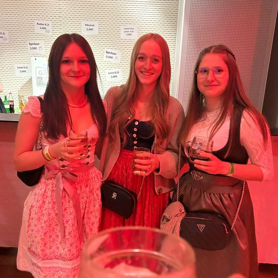 Drei Frauen in traditioneller Kleidung, die lächeln und Gläser halten, stehen zusammen unter rotem Licht.