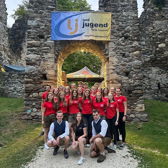 Eine Gruppe von Menschen in roten und weißen Shirts posiert für ein Foto vor einem steinernen Bogengang, mit einem Banner darüber, das Land Jugend sagt.