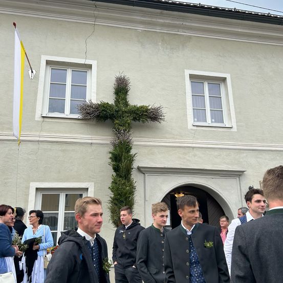 Männer in traditioneller Kleidung versammeln sich vor einer Kirche mit einem mit Grün bewachsenen Kreuz und einer Fahne.