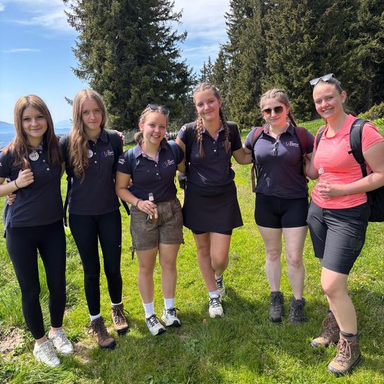 Fünf junge Frauen in Uniform stehen auf einem Grasfeld. Sie lächeln und scheinen für ein Foto zu posieren. Im Hintergrund sind Bäume und ein Berg zu sehen.
