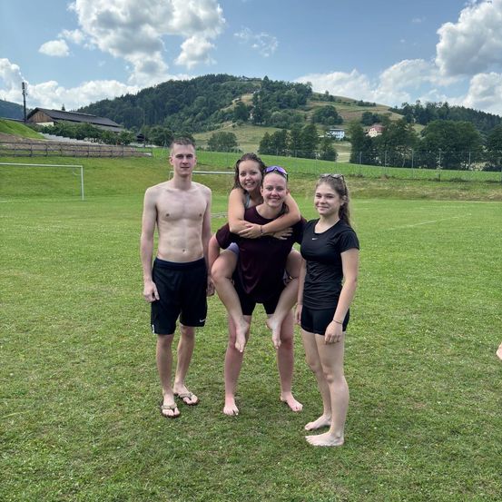 Eine Gruppe von Menschen steht auf einem Rasenfeld und posiert für ein Foto. Eine Frau hält ein Kind auf ihren Schultern, während eine andere Frau neben ihr steht. Ein Mann steht hinter ihnen. Dahinter befindet sich ein Rasenfeld mit Zaun, Tor und Bäumen. In der Ferne befindet sich ein Berg mit einem Turm.