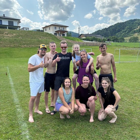 Eine Gruppe junger Leute posiert für ein Foto auf einem Rasenfeld mit einem Fußballtor im Hintergrund. Sie lächeln und machen Daumen hoch.