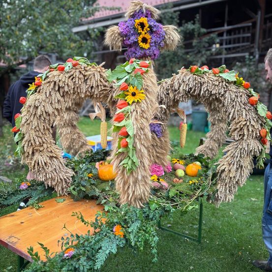Ein großer dekorativer Kranz aus Weizen und Blumen steht auf einem Holztisch, umgeben von Kürbissen und anderem Gemüse. Menschen stehen dahinter.