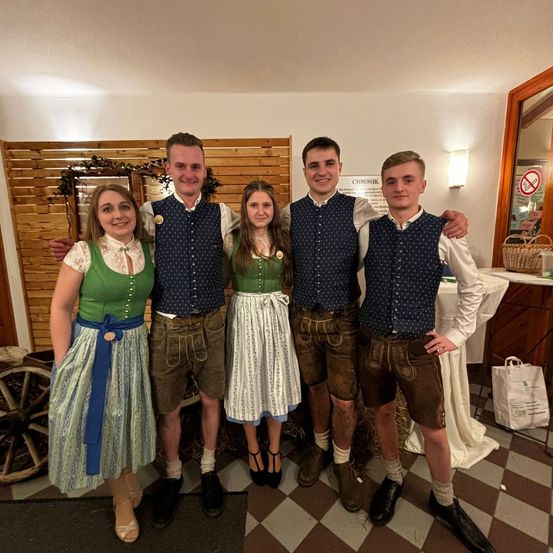 Fünf Personen in traditioneller deutscher Kleidung posieren für ein Foto in einem Raum mit Holzwänden und gemustertem Boden. Ein Holzrad und Körbe sind im Hintergrund sichtbar.