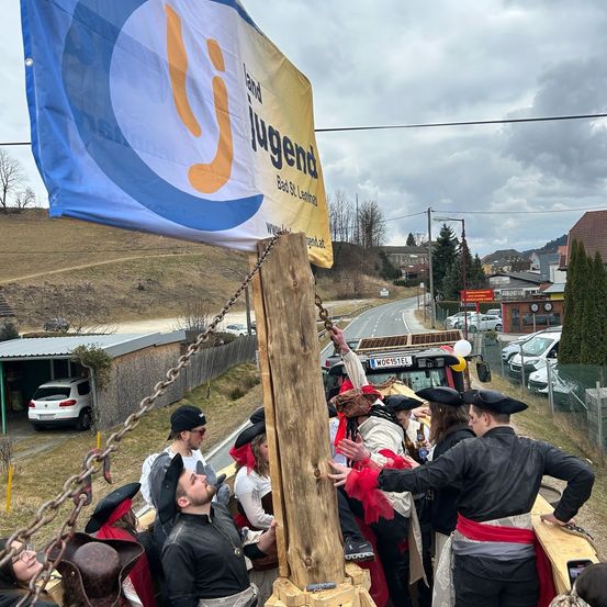 Eine Gruppe von Menschen in Piratenkostümen reitet auf einer Holzplattform, die an einen Lastwagen mit einer Flagge angeschlossen ist. Sie sind auf einer Straße, mit geparkten Autos und einem Haus in der Nähe. Ein Berg ist in der Ferne.