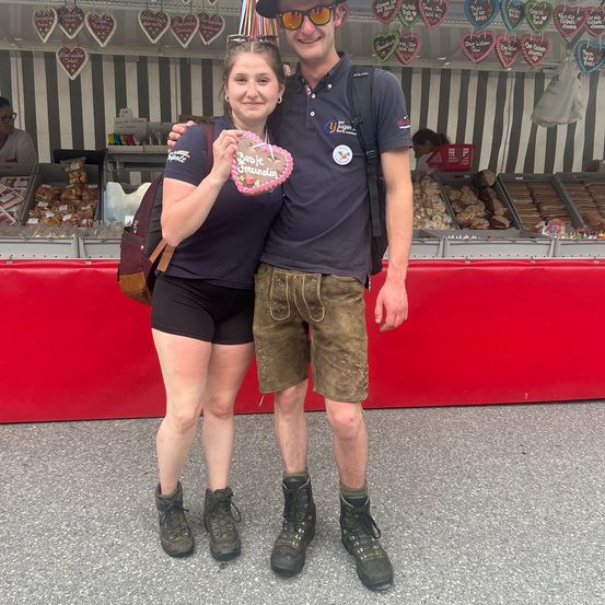 Ein Paar steht vor einem Stand, kleidet sich in traditioneller deutscher Kleidung, wobei einer ein herzförmiges Cookie hält.