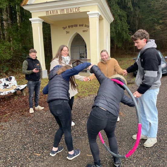 Fünf Menschen spielen draußen vor einer Kapelle mit einem Hula-Hoop. Eine Frau mit Brille lächelt.