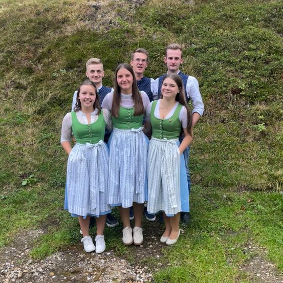 Fünf junge Leute posieren für ein Foto in traditioneller Kleidung, zwei Mädchen in grünen Kleidern und drei Jungen in Freizeitkleidung auf einem grasbewachsenen Hügel.