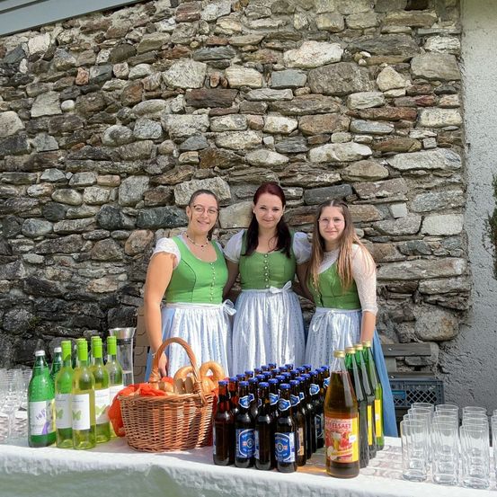 Drei Frauen in grünen Kleidern posieren hinter einem Tisch voller verschiedener Getränke, darunter Flaschen Bier und Wein, und einem Korb mit Brezeln.