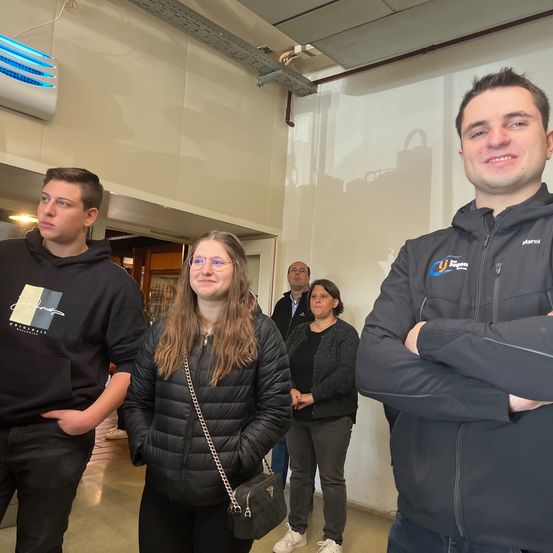 Vier Personen stehen lächelnd in einem Flur. Ein Mann und eine Frau vorne, eine weitere Frau dahinter und ein Mann im Hintergrund. Eine weiße Klimaanlage ist an der Decke.