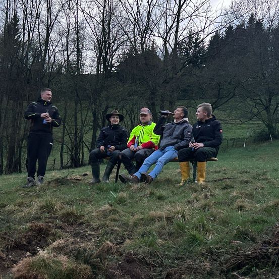 Fünf Männer sitzen auf einer Bank in einem Feld mit Bäumen und trinken aus Flaschen. Ein Mann steht und trinkt aus einer Flasche.