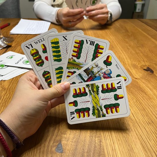 Zwei Personen sitzen an einem Holztisch und halten ein Set Tarotkarten. Eine Person hält die Karten, während die andere sie untersucht.