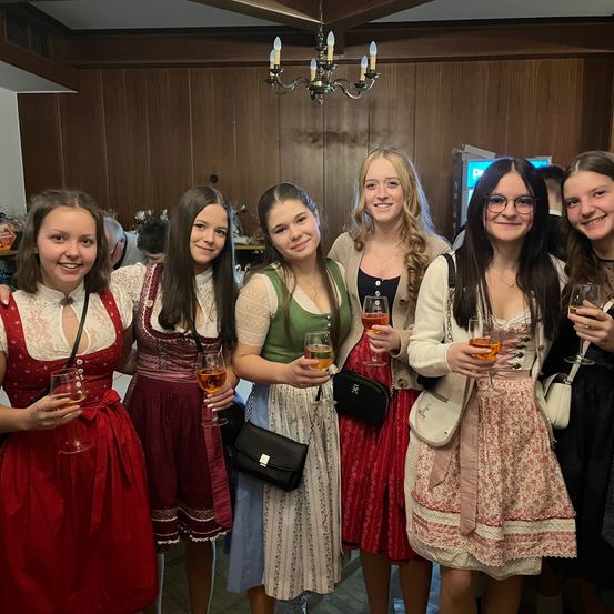 Eine Gruppe von Mädchen in traditioneller deutscher Kleidung posiert mit Weingläsern und lächelt nach vorne. Sie befinden sich in einem Raum mit Holzwänden und einem Kronleuchter.