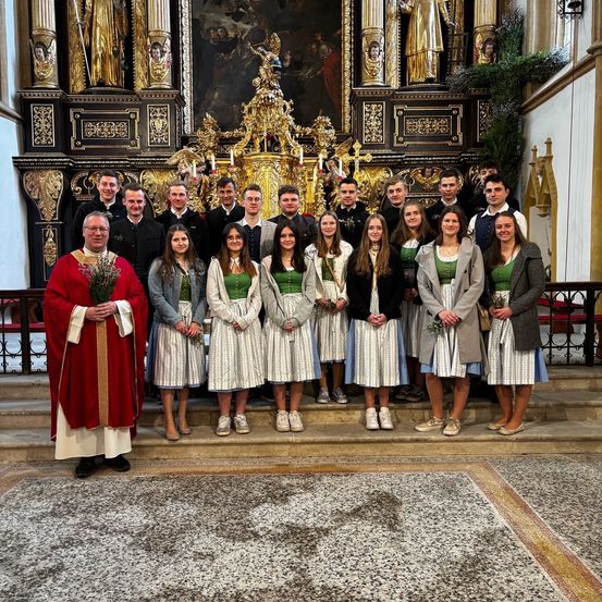 Ein Priester und eine Gruppe junger Menschen in traditioneller Kleidung posieren in einer Kirche. Sie stehen auf Stufen und halten Blumen, mit einem goldenen Altar dahinter.