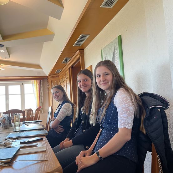 Drei junge Frauen sitzen lächelnd an einem Esstisch. Die Frau rechts trägt eine Uhr und eine Halskette. Auf dem Tisch befinden sich ein Buch, ein Stift und Besteck. Hinter ihnen ist eine Wand mit einem Bilderrahmen.