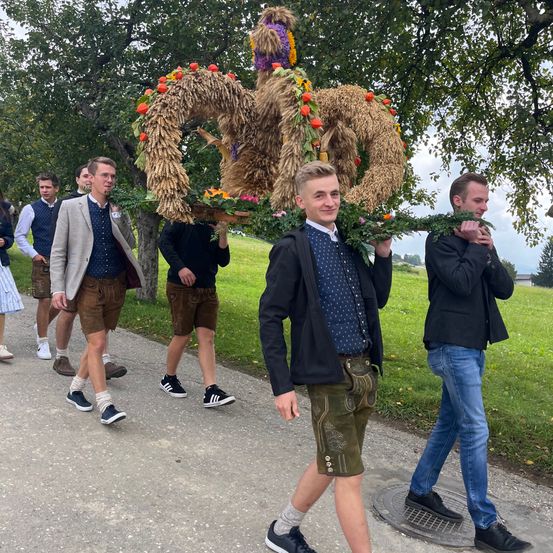 Eine Gruppe von Männern in traditioneller Kleidung geht auf einer Straße, einer von ihnen trägt eine dekorative Girlande. Bäume und Gras befinden sich auf beiden Seiten.