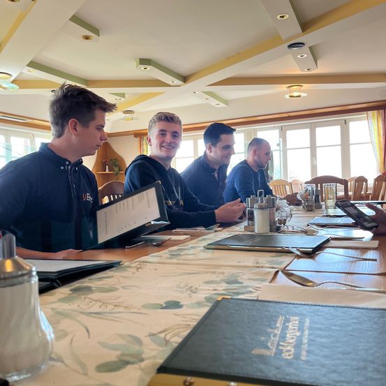 Vier Männer sitzen an einem Tisch in einem Restaurant und schauen sich die Speisekarten an. Ein blumiger Tischläufer, Salzstreuer und ein Handy sind auf dem Tisch. Der Raum hat Glasfenster, und es gibt Stühle und einen Holzschrank.