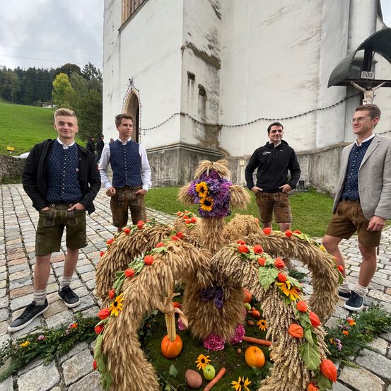 Vier junge Männer in traditioneller bayerischer Kleidung stehen vor einer Kirche. Sie lächeln und posieren für ein Foto. Ein großer Blumenschmuck ist im Vordergrund.