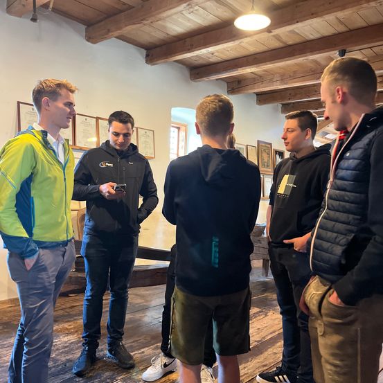 Fünf Männer stehen in einem Raum. Einer hält ein Handy. Ein anderer trägt eine neongrüne Jacke. Der Raum hat Holzbalken und gerahmte Bilder an der Wand.