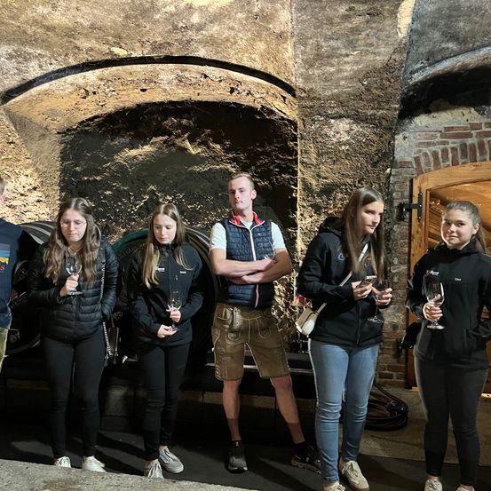 Eine Gruppe junger Menschen steht in einem Keller und hält Weinläser in den Händen. Sie lächeln und scheinen für ein Foto zu posieren.