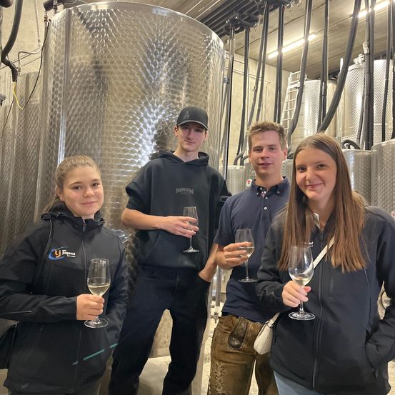 Drei Personen posieren für ein Foto in einer Fabrik. Sie halten Weinglas und zwei lächeln. Im Hintergrund sind Metalltanks und Rohre zu sehen.