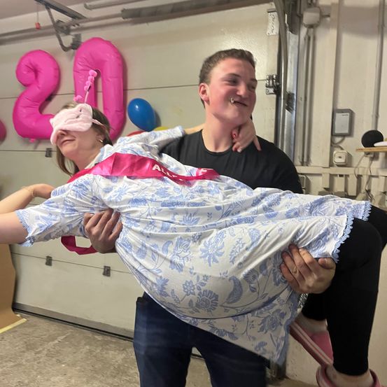 Eine Frau mit einer Augenbinde und einem Blumenkleid wird von einem Mann in einer Garage angehoben, mit Luftballons und einer Nummer-20-Dekoration im Hintergrund.