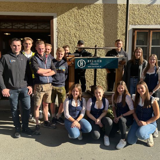 Eine Gruppe von Studenten posiert vor einem Gebäude. Einige knien, andere stehen, mit einem Hartlieb-Schild dahinter. Die Umgebung ist eine Mischung aus urbanen und ländlichen Elementen.
