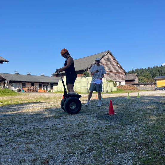 Eine Person fährt auf einem Segway um ein Feld mit Hütchen, während eine andere Person zusieht. Im Hintergrund befinden sich Gebäude und Bäume.