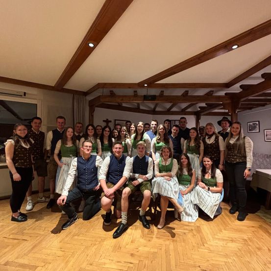 Eine Gruppe von Menschen in traditioneller Kleidung posiert für ein Foto in einem Raum mit Holzbalken, Stühlen und Tischen. Die Gruppe besteht sowohl aus Männern als auch aus Frauen.