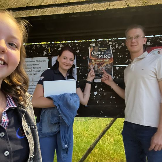 Drei Menschen stehen vor einer Holzstruktur, wobei eine Frau ein Buch und ein Poster für Castle on Fire hält. Der Mann neben ihr zeigt auf das Poster. Hinter ihnen steht ein Schild und ein Grasfeld.