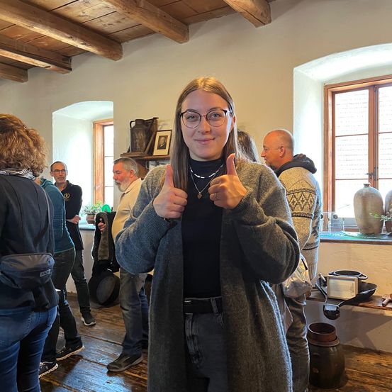 Eine Frau in einem grauen Cardigan gibt zwei Daumen hoch in einem Raum mit Holzböden und Balken. Andere Menschen stehen herum und einige Gegenstände sind auf einem Regal und Fensterbank sichtbar.