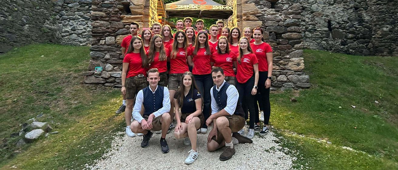 Eine Gruppe von Menschen in roten T-Shirts steht vor einem Steinbogen mit einem Banner, auf dem 'Land Jugend' steht. Sie lächeln und posieren für ein Foto.