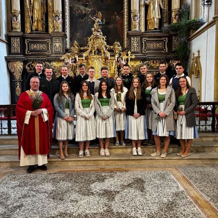 Eine Gruppe von Menschen steht vor einem Altar einer Kirche, gekleidet in traditionelle Kleidung, mit einem Priester, der Blumen hält.