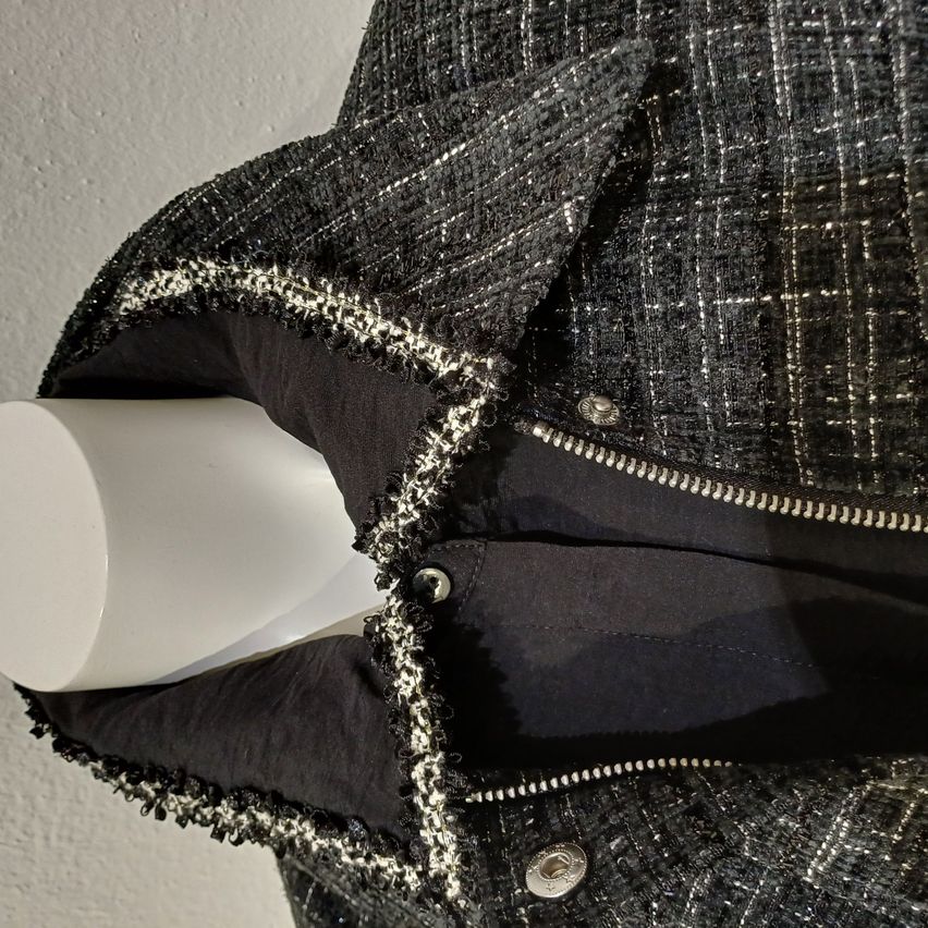 Eine Nahaufnahme eines schwarzen und grauen Tweed-Jacken mit Reißverschluss und Kapuze. Die Jacke hat eine glänzende Textur und dekorative Fransen an den Kanten.