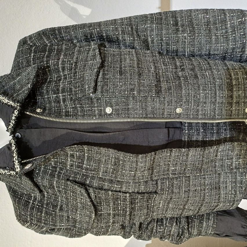 Eine schwarze und graue Tweedjacke mit Reißverschluss, zwei Knöpfen und zwei Taschen vorne. Sie ist gefaltet und auf einer weißen Oberfläche ausgestellt.