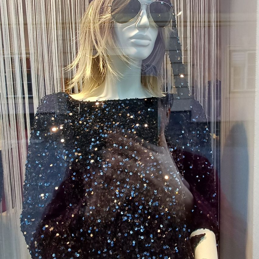 A mannequin wearing a sequined black dress with sunglasses, inside a shop window.