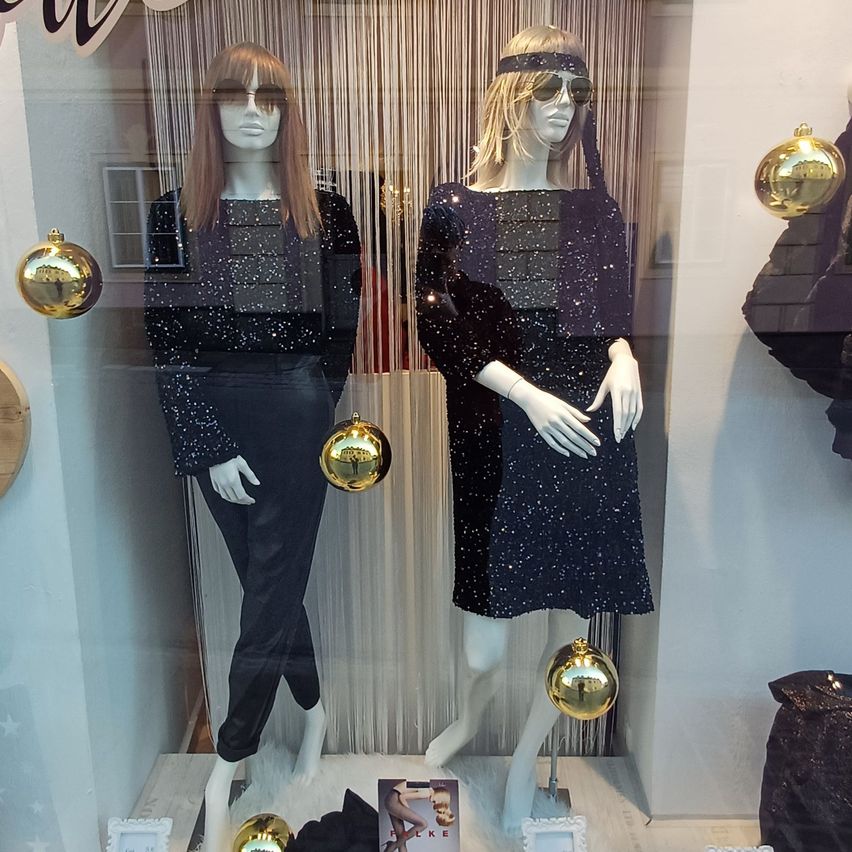 Two mannequins wearing sequined black outfits stand in a window display. They have sunglasses on and are surrounded by gold ornaments. A magazine is on the display table.