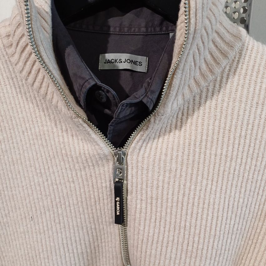 A close-up of a white, knitted jacket with a zipper and a label that reads Jack & Jones. The zipper is partially open, revealing a black inner shirt.