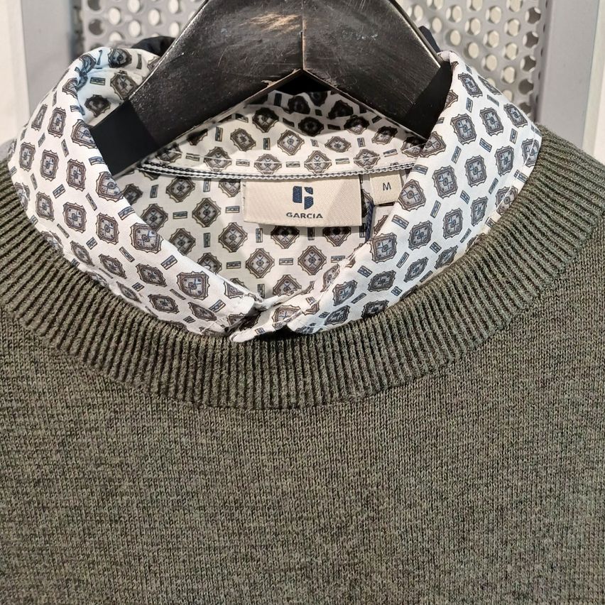 A gray sweater with a patterned collar, displayed on a hanger with a metal background. The collar features a black and white geometric design.