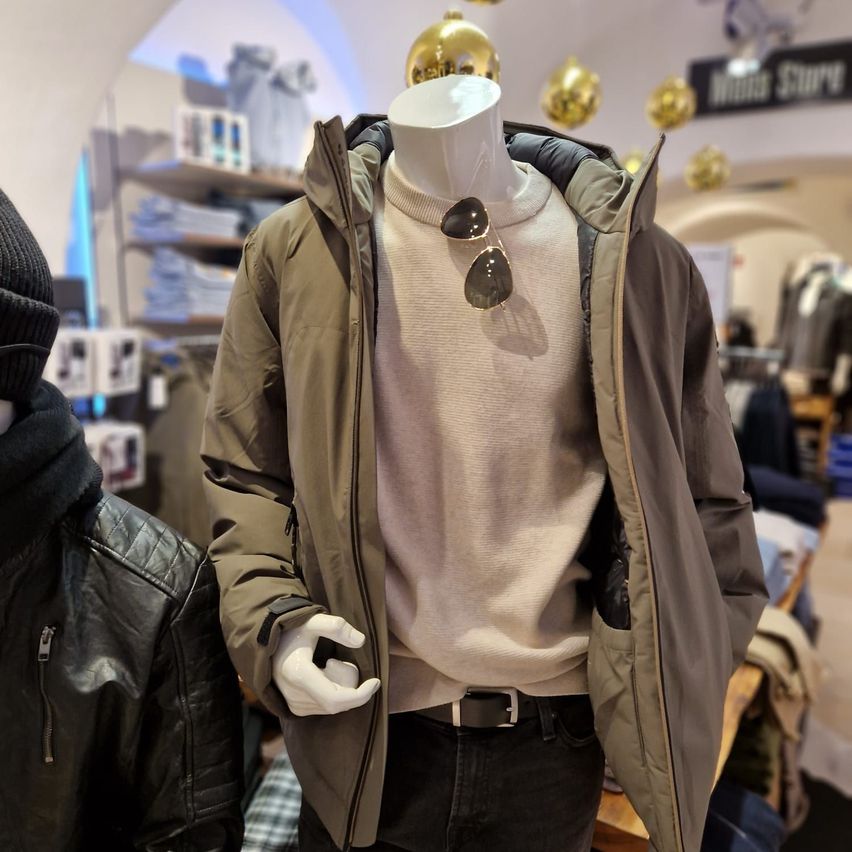Ein Mannequin trägt eine braune Jacke, ein weißes Rollkragenshirt, eine Sonnenbrille und schwarze Hose in einem Bekleidungsgeschäft.
