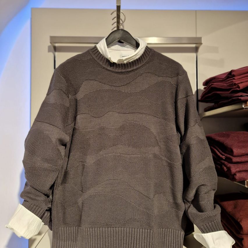 Ein graues Sweatshirt mit strukturiertem Muster hängt an einem Kleiderbügel in einem Bekleidungsgeschäft. Dahinter sind weitere gefaltete Kleidungsstücke zu sehen.