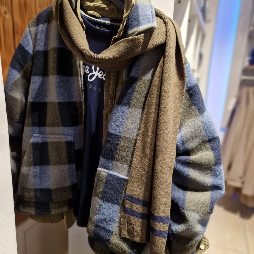 Eine blau-weiß karierte Jacke mit einem braunen Schal wird in einem Geschäft ausgestellt. Die Jacke hat Taschen und einen Reißverschluss. Der Schal ist ordentlich gefaltet.