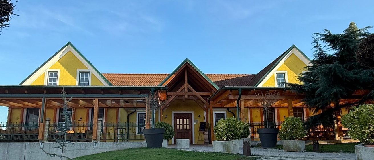 Ein gelbes Gebäude mit einem Ziegeldach und einer Holzveranda. Es hat mehrere Fenster und ist von Topfpflanzen umgeben. Ein Ziegelweg führt zum Eingang.