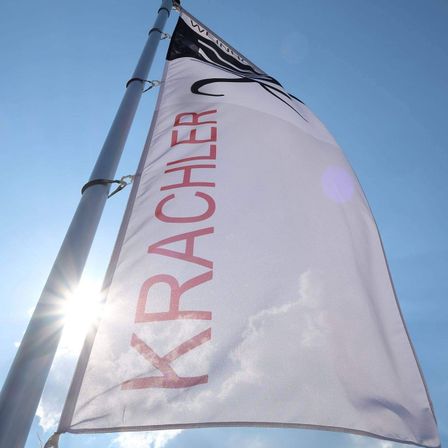Ein weißes Banner mit rotem Text, der 'Krachler' lautet, flattert im Wind gegen einen blauen Himmel, teilweise von Sonnenlicht beleuchtet.