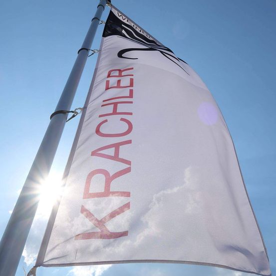 Ein weißes Banner mit rotem Text, der 'Krachler' lautet, flattert im Wind gegen einen blauen Himmel, teilweise von Sonnenlicht beleuchtet.