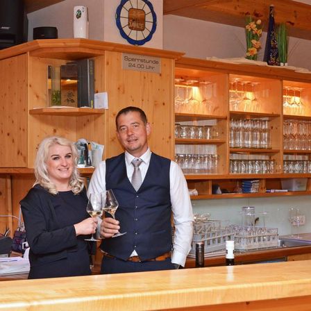 Ein Mann und eine Frau stehen hinter einer Bar, halten Weingläser in der Hand und lächeln. Hinter ihnen befinden sich Regale mit Weingläsern und ein Schild mit der Aufschrift 'Sperrstunde 24.00 Uhr'.